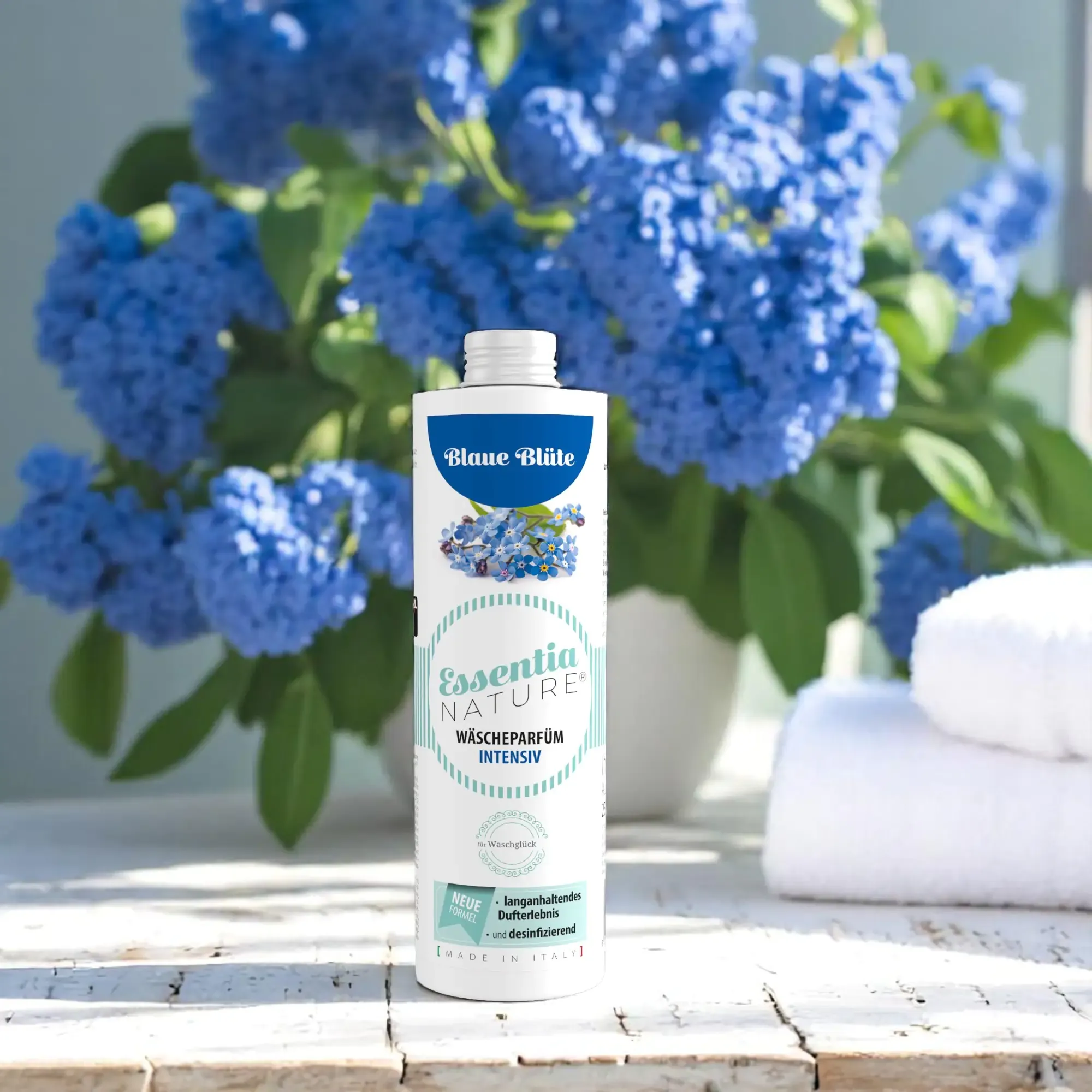 Essentia Nature wasparfum Intensiv Blaue Blüte van Wasgeluk, microplasticvrij wasparfum met langdurige frisse bloemengeur, afgebeeld op een witte houten tafel met blauwe bloemen en stapels zachte, witte handdoeken.