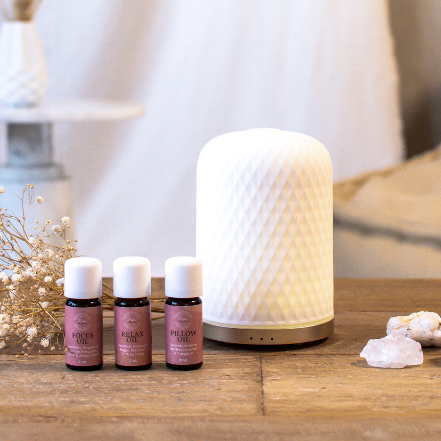 Stijlvolle witte aroma-diffuser met drie roze Wasgeluk geurolieën (Focus Oil, Relax Oil en Pillow Oil) op een houten tafel in een warme, rustgevende setting voor een langdurige, luxe geurbeleving in huis.