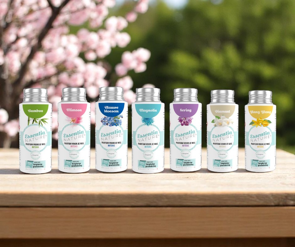 Rij Essentia Nature wasparfum flesjes in de geuren Bamboo, Mimosa, Blauwe Bloesem, Magnolia, Sering, Diamant en Ylang Ylang op een houten tafel in een groene, bloemrijke tuin voor een langdurige, luxe en microplasticvrije geurbeleving van Wasgeluk.