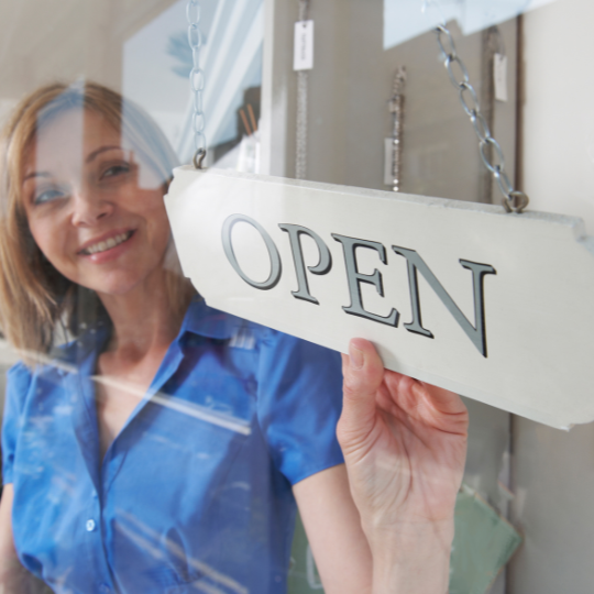 Lachende vrouw in een blauwe blouse hangt een wit bord met de tekst “OPEN” aan de deur van de Wasgeluk-winkel met wasparfum en duurzame, microplasticvrije wasproducten voor een langdurige, luxe geurbeleving.