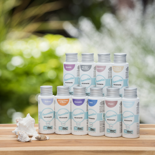 Set Essentia Wasgeluk wasparfum-concentraten in diverse geuren zoals Jasmin, Orangeblüte & Zitrone en Argon, in pastelkleurige flesjes op een houten tafel met schelpen, tegen een groene natuurlijke achtergrond voor een luxe, langdurige wasgeurbeleving.