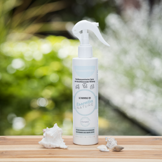 Essentia Nature Le Nuvole di desinfecterende spray van Wasgeluk in een witte fles met trigger-spray, gefotografeerd op een houten tafel met schelpen en groene, natuurlijke achtergrond voor een frisse, duurzame was- en geurbeleving.